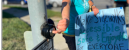 mode shift omaha member pressing a walk button or beg button and holding a sign that reads: Make sidewalks accessible, safe & enjoyable for everyone